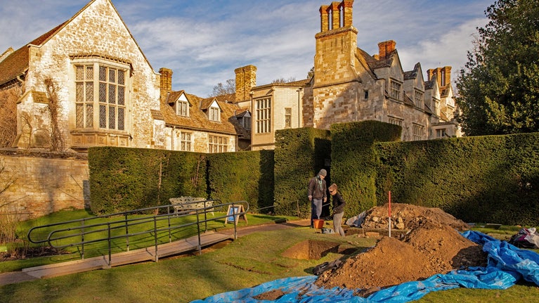 Two archaeologists stand in the corner of the Rose Garden at Anglesey Abbey looking into a test pit, surrounded by blue covers and piles of soil. The house is lit up by the sunshine in the background. Blue skies.